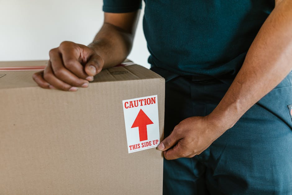 Close-up of a person during a home relocation, holding a cardboard box with a red and white caution label indicating 'This Side Up' and an upward arrow. The individual’s hands are gripping the box firmly, with part of their arms and torso visible, dressed in casual clothing suited for packing and moving tasks. The box appears to be made of sturdy cardboard, and the caution label signifies careful handling during the furniture transport process. The background suggests an indoor environment, likely a room within a house or apartment, where packing materials and other boxes may be present for the systematic packing and loading process required for house removals. The image captures the careful handling and organization involved in the loading phase of a professional move, which [COMPANY_NAME], such as Big Man, may assist with as part of their removals services, supporting efficient and secure house relocation with proper packing and transportation practices.