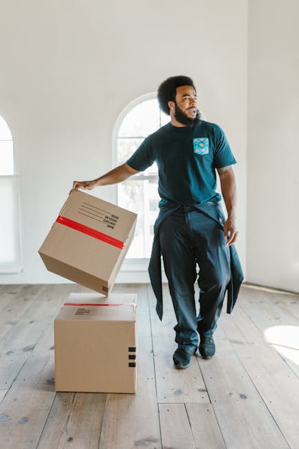 Close-up of a person during a home relocation, holding a cardboard box with a red and white caution label indicating 'This Side Up' and an upward arrow. The individual’s hands are gripping the box firmly, with part of their arms and torso visible, dressed in casual clothing suited for packing and moving tasks. The box appears to be made of sturdy cardboard, and the caution label signifies careful handling during the furniture transport process. The background suggests an indoor environment, likely a room within a house or apartment, where packing materials and other boxes may be present for the systematic packing and loading process required for house removals. The image captures the careful handling and organization involved in the loading phase of a professional move, which [COMPANY_NAME], such as Big Man, may assist with as part of their removals services, supporting efficient and secure house relocation with proper packing and transportation practices.