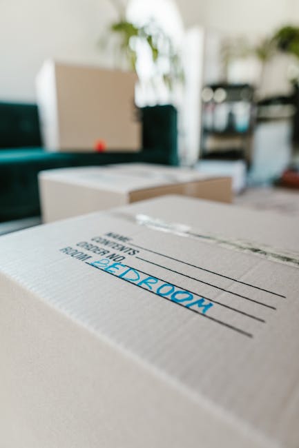 A close-up view of a cardboard moving box labeled 'BEDROOM' with blue writing, situated indoors in a home environment. In the background, additional packed boxes are visible, along with household furniture such as a sofa, a coffee table, and shelving units. The boxes are wrapped in clear packaging tape and are stacked or placed on the floor near a doorway and a large window, allowing natural light to illuminate the scene. The setting reflects the packing stage of a home relocation or furniture transport process, with some boxes appearing opened or partially filled. This image illustrates the packing and moving process typically undertaken during a house removal, highlighting the importance of organized packing and preparation before a professional removal service like Big Man, which specialises in removals, helps facilitate a stress-free house move.