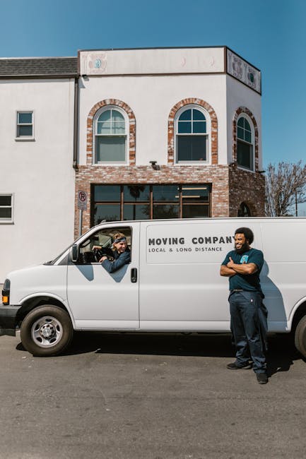 The image shows a white van labeled 'MOVING COMPANY LOCAL & LONG DISTANCE' parked on a street in front of a two-storey residential building with brick and white exterior walls. Inside the van's cab, a person is visible reaching out the window, engaging in conversation or handover with a resident. Outside, a mover in dark clothing stands with arms crossed, observing the process, indicating a home relocation or furniture transport activity. The scene takes place during daylight hours with a clear blue sky. The building has two arched windows on the upper floor and a large ground-floor window, suggesting a suburban or urban setting. The image illustrates aspects of packing and moving logistics typical of professional removals services, relating to house or apartment moves, and emphasizes the importance of proper loading and communication during furniture transport, supported by the presence of moving equipment such as the van and the actions captured in the scene.