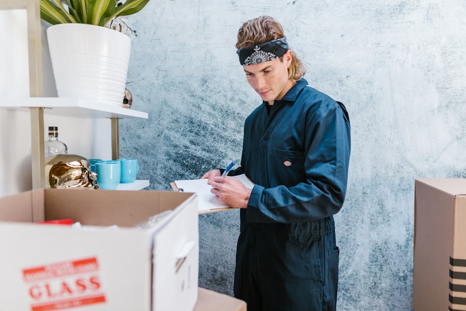 A young male mover dressed in dark blue overalls and a black bandana with white patterns is inside a room, standing next to a cardboard box filled with packed items. He is holding a clipboard and a pen, likely reviewing a packing list or inventory as part of a home relocation process. Behind him, there is a white shelving unit with decorative items, including a potted plant in a white ceramic pot and golden ornaments, alongside blue cups and small bottles. The room has a textured, light-colored wall, and the scene is well-lit. The presence of packed boxes and the mover's activity suggests an organized packing and moving operation, typical of professional removals carried out by companies like Big Man, which offers services in furniture transport and loading during house removals.